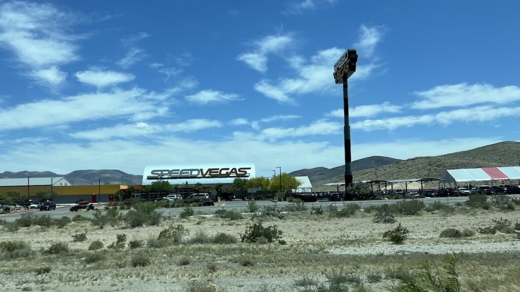 Photo of a distant view of the Speedstyle facilities and its big sign from the nearby freeway. Mountains in the background, race track middle ground, and scrub desert in the foreground.